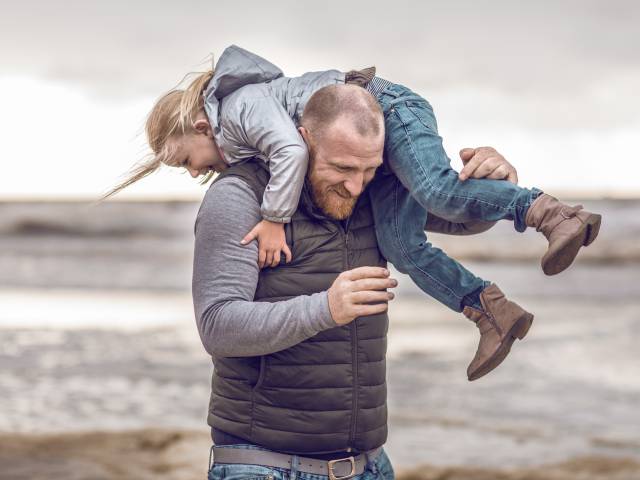 Papa trägt Tochter über der Schulter mit der Nordsee im Hintergrund