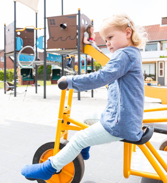 Mädchen auf einem Dreirad und andere Kinder spielen auf dem Spielplatz