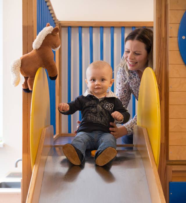 Kleiner Junge sitzt auf einer Kinder rutsche und die Mutter hält ihn 