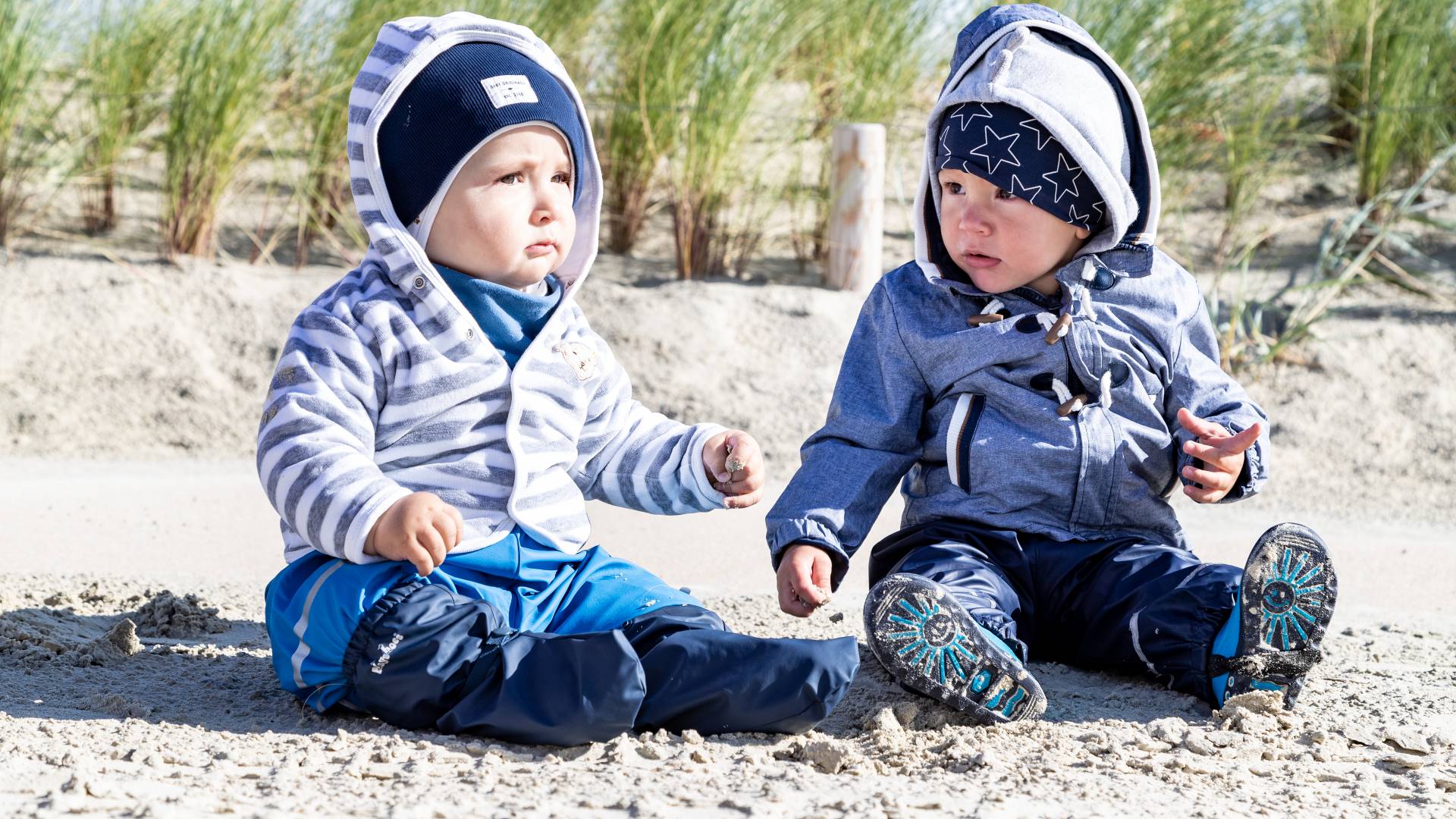 Zwei kleine Kinder sitzen warm eingepackt am Strand der Nordsee