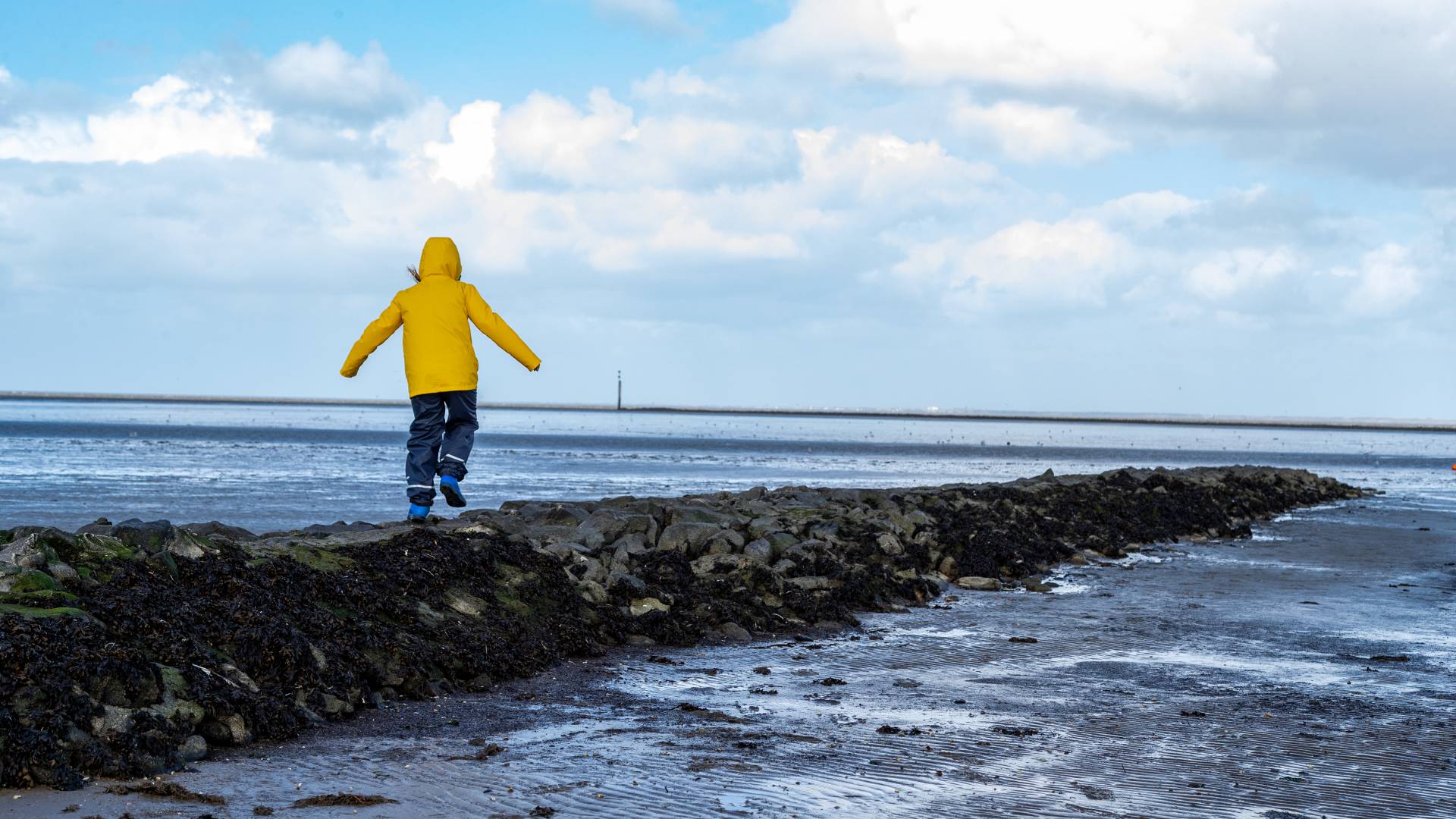 Kind mit gelber Jacke läuft über Steine an der Nordsee