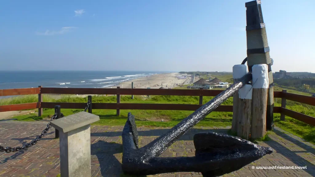 Ostfriesland mit dem Boot erkunden | Großes Anker-Denkmal auf Norderney, welches über eine Bootstour erreichbar ist , mit Blick auf den Strand, klaren blauen Himmel und Wellen der Nordsee, Strandkörbe und Gebäude im Hintergrund 